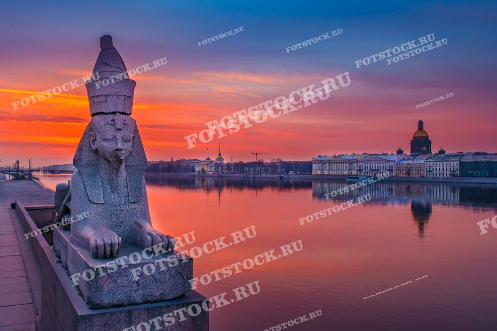 На фотографии изображён сфинкс на набережной Невы в Санкт-Петербурге на фоне яркого заката. Вдалеке виден купол Исаакиевского собора и здания Адмиралтейства. Отражение небесных красок на воде создаёт живописный вид. Это изображение идеально подходит для использования в туристических путеводителях, рекламных материалах, посвящённых Санкт-Петербургу, а также в образовательных и культурных публикациях.